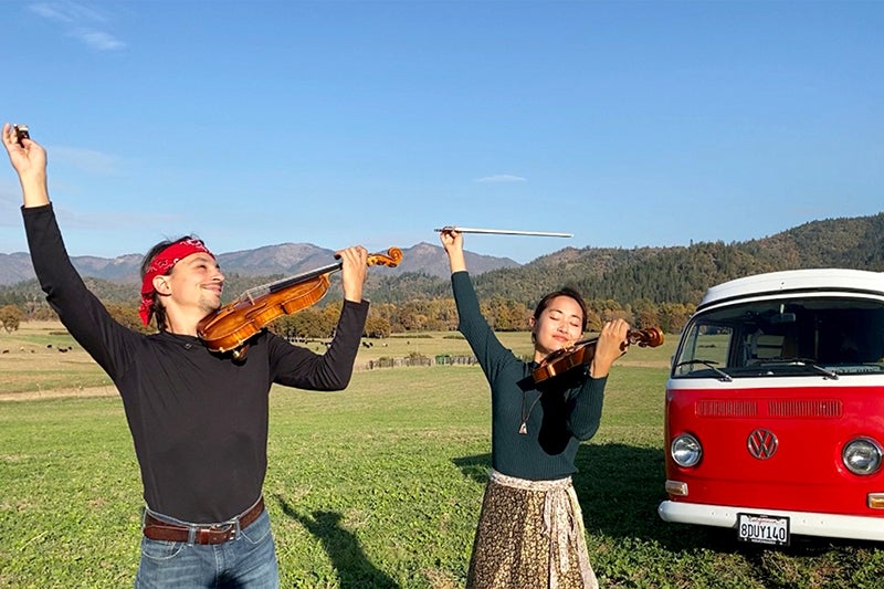 Man and woman playing violins with a van in the background