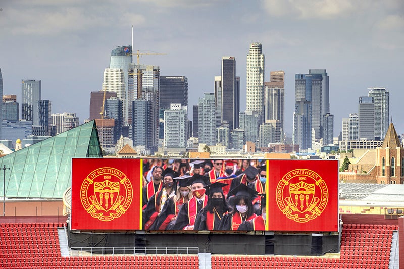 An image of the LA Coliseum big screen showing USC's graduation, with the Downtown Los Angeles skyline in the backdrop